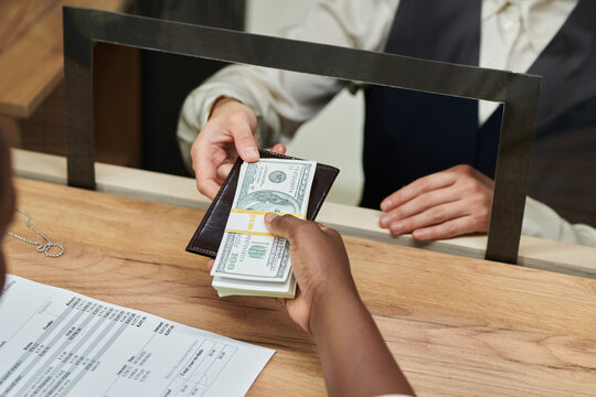Black adult hand holding wallet and stack of dollar bills, passing money to Caucasian adult bank teller behind counter, financial transaction occurring at service window