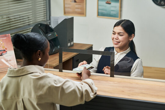 Caucasian young adult woman handing cash to Black young adult woman across counter in bank office, both women engaging in financial transaction, visible office equipment in background