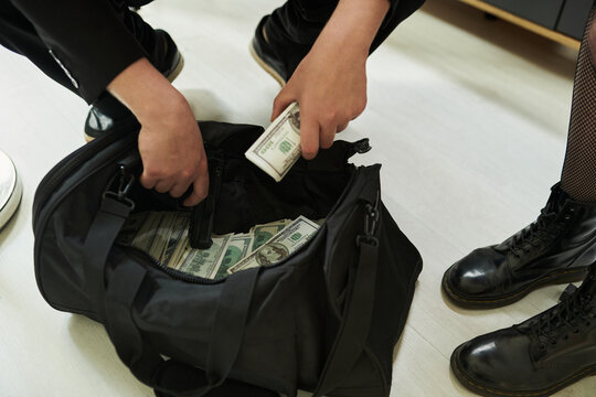 Unrecognizable man placing bundles of cash into black duffel bag next to woman wearing boots standing nearby