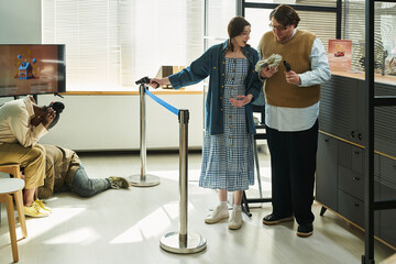 Caucasian man holding stack of cash standing next to woman smiling during bank robbery, victims with heads in hands behind barrier