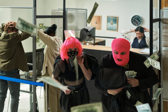 Two masked young adults wearing black clothing robbing bank, holding bags and cash while multiethnic bank employees and customers standing behind with hands on heads, money flying in air