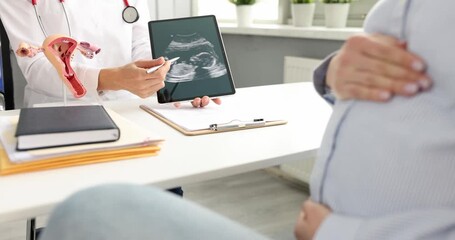Female doctor shows ultrasound scan on tablet to pregnant woman. Professional explains fetus position and points at screen during appointment