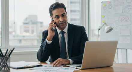 Business man communicating while working at modern office desk, focused professional discussing tasks on phone or video call, multitasking with laptop, corporate communication and productivity concept