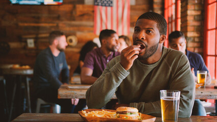 Man Watching Game On Screen In Sports Bar Eating Burger And Fries