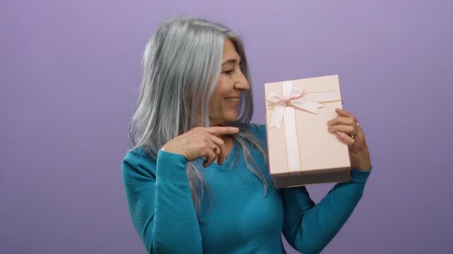 Senior woman with grey hair in a blue sweater looks delightedly at a pink gift box with a ribbon against a purple background, showcasing surprise and joy.