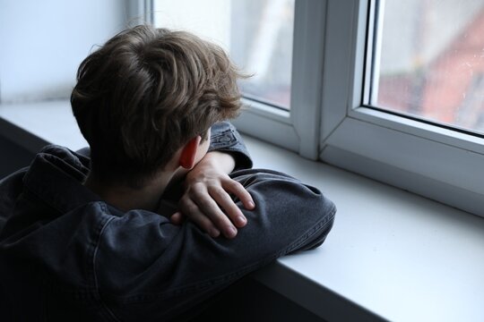 Depressed teenage boy near window at home, space for text