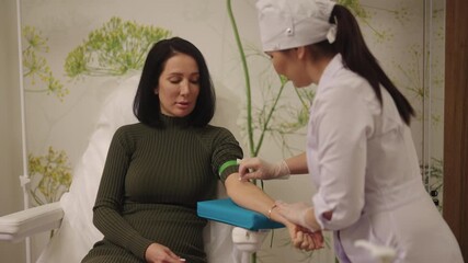 Female patient sits in clinic chair while nurse in gloves disinfects her arm and applies tourniquet preparing for blood draw for laboratory testing, health check or donation. Shooting in slow motion.
