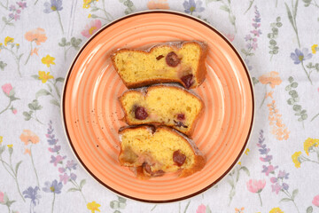Three slices of baked cherry cake dusted with powdered sugar are stacked vertically on a patterned orange plate against a floral tablecloth background