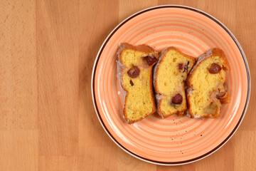 Three rectangular slices of baked yellow cake with cherries, dusted with powdered sugar, are arranged on a patterned orange plate on a wooden table