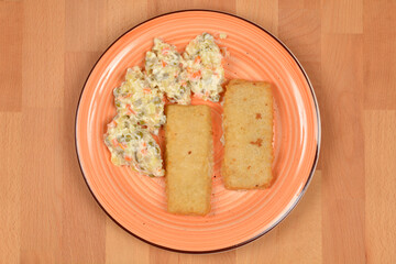 Two rectangular breaded fish filets served with a portion of classic French salad (Croatian way) on an orange-rimmed plate on a light wood surface.