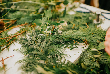 A table covered in green leaves and twigs. A person is working on a wreath made of these leaves