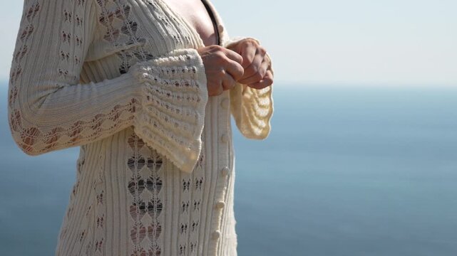 Woman, unbuttoning, cardigan, female unbuttons white knitted cover-up over bikini with ocean backdrop on a sunny day