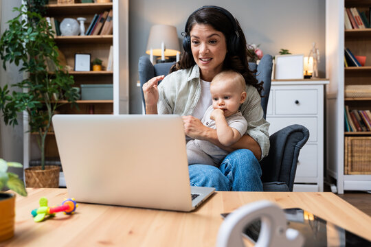 Working mom on video call with headphones holding her baby and waving to colleagues balancing parenting and remote work staying professional managing distractions, keeping communication warm from home