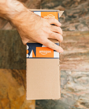 Paris, France - Dec 13, 2024: Close-up of a hand holding a cardboard Amazon delivery box sealed with branded orange and navy tape, photographed indoors on a stone floor with natural warm lighting.