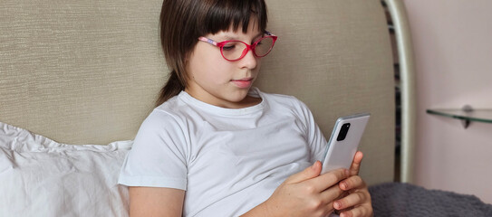 Child teenager girl in glasses reading on smartphone sitting on bed at home