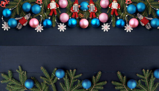 A frame of fir branches, blue and pink Christmas tree baubles, and Christmas doll figurines on a blue wooden background.