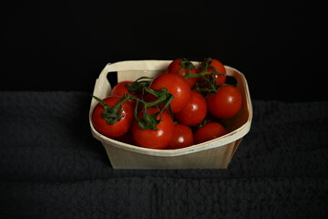 Fresh ripe cherry tomatoes in a small wooden basket on a dark fabric background. Minimalist still life with natural light and organic feel. Concept of healthy eating, simplicity, and slow living.