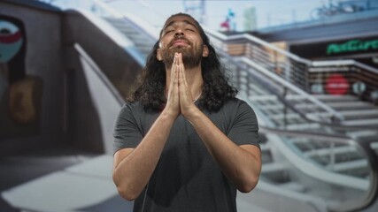 Man pressing bare hands together praying by an escalator inside a shopping mall building, eyes closed and head tilted back; gratitude prayer.