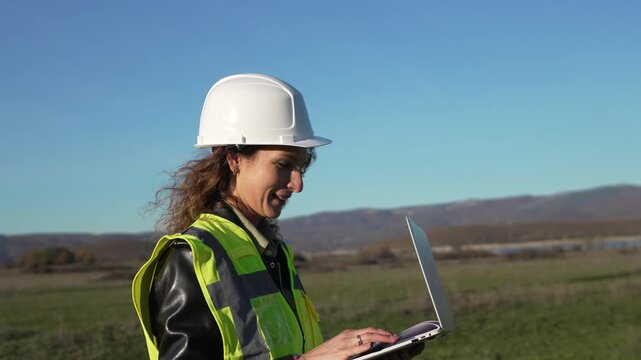 Engineer surveyor woman checking land site with laptop computer wearing hardhat and safety vest outdoors.