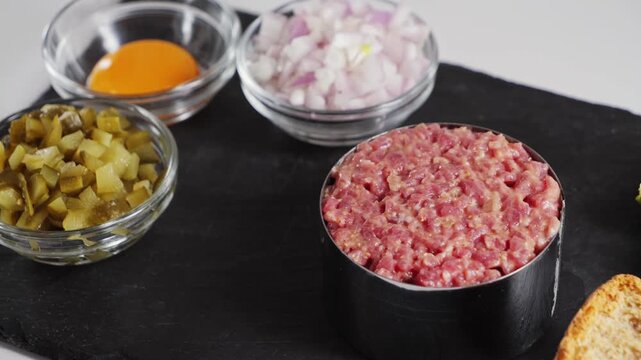 Raw beef tartare steak with ingredients for prepare, chopped onion, capers, yolk. Close up of cooking a delicious French dish in a kitchen. High quality 4k footage