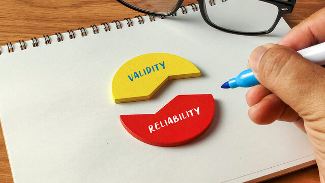 Hand writing next to Validity and Reliability wooden puzzle pieces on a notebook with glasses for research and quality concepts.