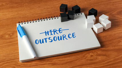 The word Hire crossed out and Outsource written on a notebook with blocks on a wooden desk...
