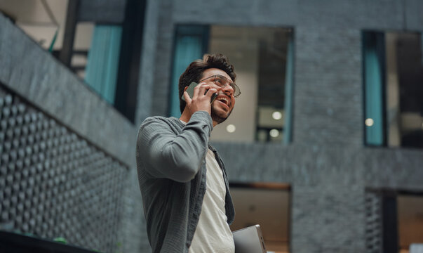 diverse male entrepreneur talking on phone in modern atrium, looking upward, wearing glasses and casual