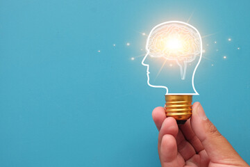 Creative idea and innovation concept. Hand holding lightbulb with glowing brain in head silhouette. Brainstorming, AI, knowledge, and new solution on blue background with copy space.