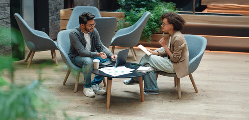 colleagues meeting in modern lounge, planning strategy over documents and coffee, reviewing laptop graphs