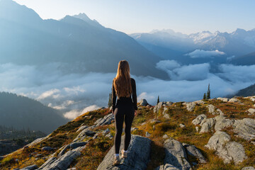 A woman stands on a peak, gazing at a stunning mountain vista with clouds.
