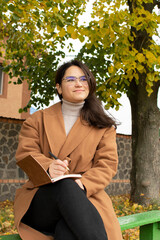 Young attractive woman in brown coat sitting on bench and writing in notebook	