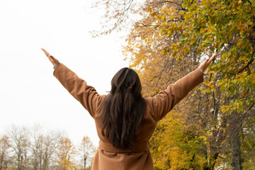 A woman wearing a brown coat stands with her arms raised under autumn trees