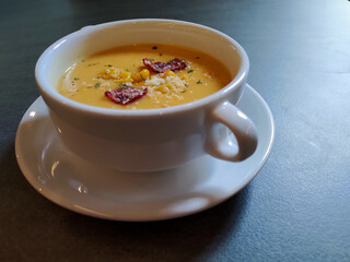 A white bowl of creamy yellow soup topped with corn, small meat slices, herbs, and grated cheese, placed on a gray table.