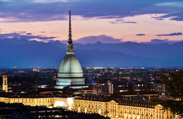 Night skyline of Turin or Torino, Italy showcasing the iconic Mole Antonelliana illuminated against a backdrop of the Alps and colorful twilight sky, with the city lights glowing warmly below