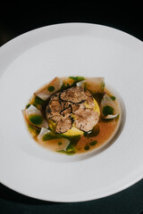 A white plate with a dish of food on it. The dish is a combination of meat and vegetables, with a sauce on top. The plate is placed on a table, and the overall mood of the image is appetizing