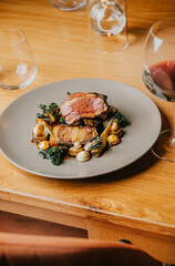 A plate of food with meat and vegetables on a wooden table. The plate is placed on a wooden table with a chair next to it. There are wine glasses on the table as well