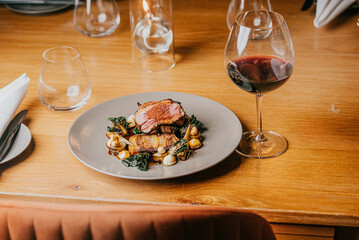 A plate of food with a glass of red wine on a wooden table. The plate has meat and vegetables, and the wine glass is half full. The scene is set in a restaurant, and the atmosphere is elegant