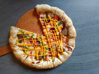 Close-up of a baked pizza topped with cheese, sliced meat, green peppers, and drizzled with yellow sauce on a gray surface.