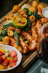 A plate of food with shrimp, tomatoes, and a lemon wedge. The plate is on a wooden board and is accompanied by a glass of wine