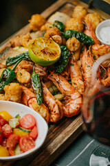 A plate of food with shrimp, tomatoes, and a lemon wedge. The plate is on a wooden board and is accompanied by a glass of wine
