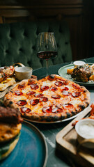 A pizza with pepperoni sits on a table next to a wine glass. The table is set with plates of food and a bowl of dipping sauce. Scene is casual and inviting