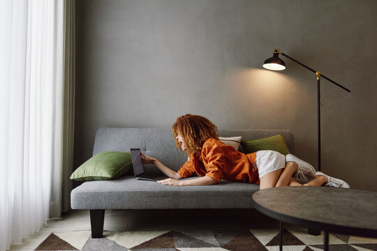 Young woman with curly hair using a laptop while relaxing on a couch in a cozy living room with modern decor and natural light, illustrating remote work and comfort