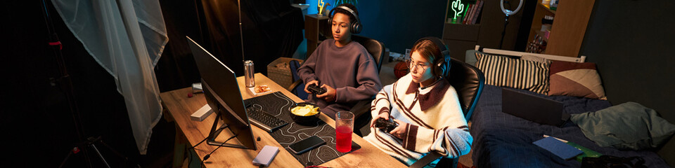 Two teenagers, Black boy and Caucasian girl, sitting at desk playing video games with controllers,...