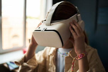Teenage Caucasian girl wearing virtual reality headset, holding device with both hands, sitting indoors near window, engaging with immersive digital experience, face partially obscured