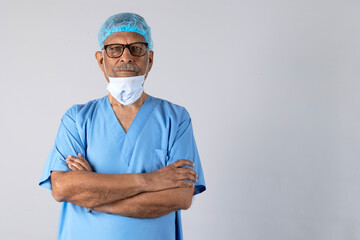 Portrait of a indian senior physiotherapist arm crossed in light blue gown and face mask deep thinking isolated studio background. Protect yourself and others medical advice against the covid-19