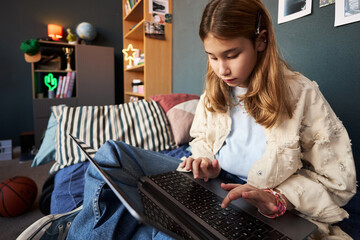 Teenage Caucasian girl sitting on bed using laptop computer, focusing on screen and typing with both hands, basketball and books visible in background, casual home environment