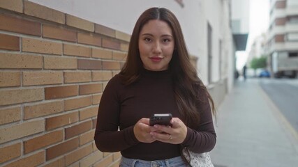 Hispanic woman scrolls smartphone on city street by brick wall while reading messages; calm contentment. - Powered by Adobe
