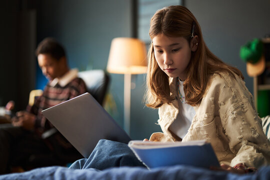 Asian teenage girl with hearing aid studying with laptop and notebook, sitting on bed, focusing on homework while Black teenage boy in background using smartphone, modern bedroom setting