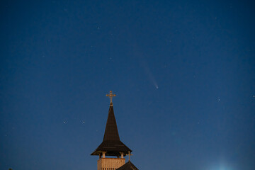 Celestial and Spiritual: Rare Comet ATLAS 2024 Shines Brightly in the Night Sky Behind the Illuminated Cross of an Old Wooden Church, Symbolizing Faith and Science