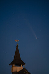 Celestial and Spiritual: Rare Comet ATLAS 2024 Shines Brightly in the Night Sky Behind the Illuminated Cross of an Old Wooden Church, Symbolizing Faith and Science